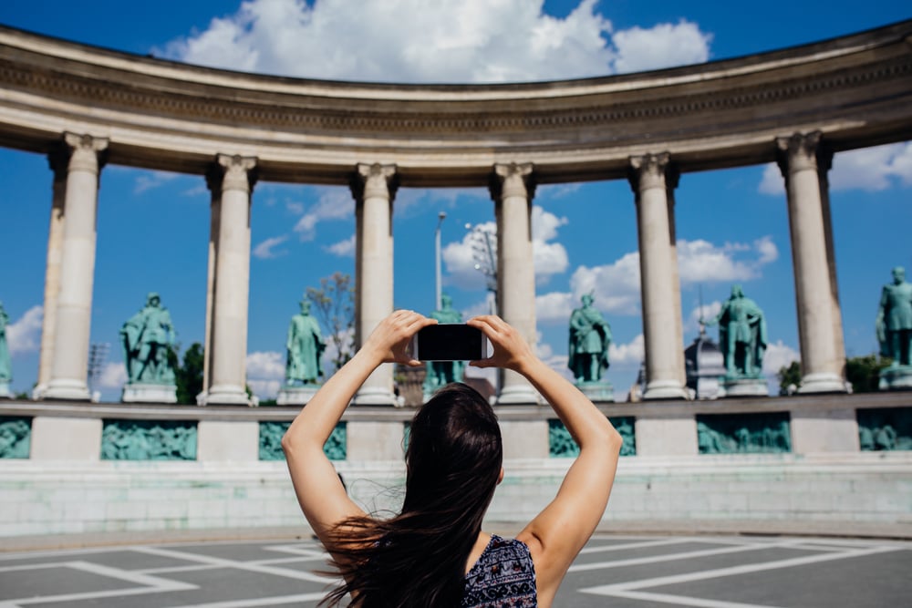 Woman tourist visiting Heroes’ Square (Hősök tere) in Budapest, photographing the Millennium Monument on Andrassy Avenue with her smartphone, a top Hungary tourism site included in the Budapest City Pass by MegaPass.