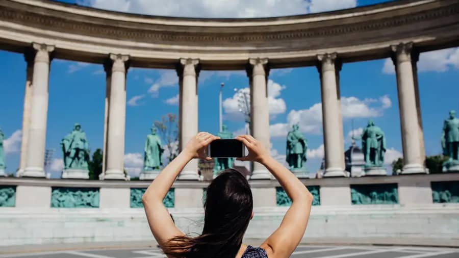 Woman tourist visiting Heroes’ Square (Hősök tere) in Budapest, photographing the Millennium Monument on Andrassy Avenue with her smartphone, a top Hungary tourism site included in the Budapest City Pass by MegaPass.